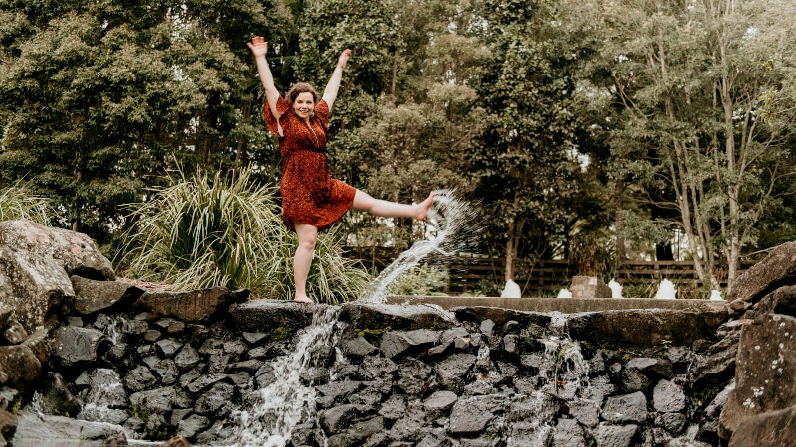 An example of authentic expression. Showing the emotion of joy. Chantelle (woman) with hands up in the air, smile on her face and one leg kicking up a stream of water into the air in front of her.